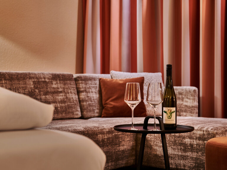Zwei Gläser Wein und eine Weinflasche stehen auf einem Tablet vor einer gemütlichen Couch